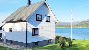 Ein zweistöckiges Haus mit einem Giebeldach, einer Fahnenmasten und Blick auf einen See und Berge im Hintergrund.