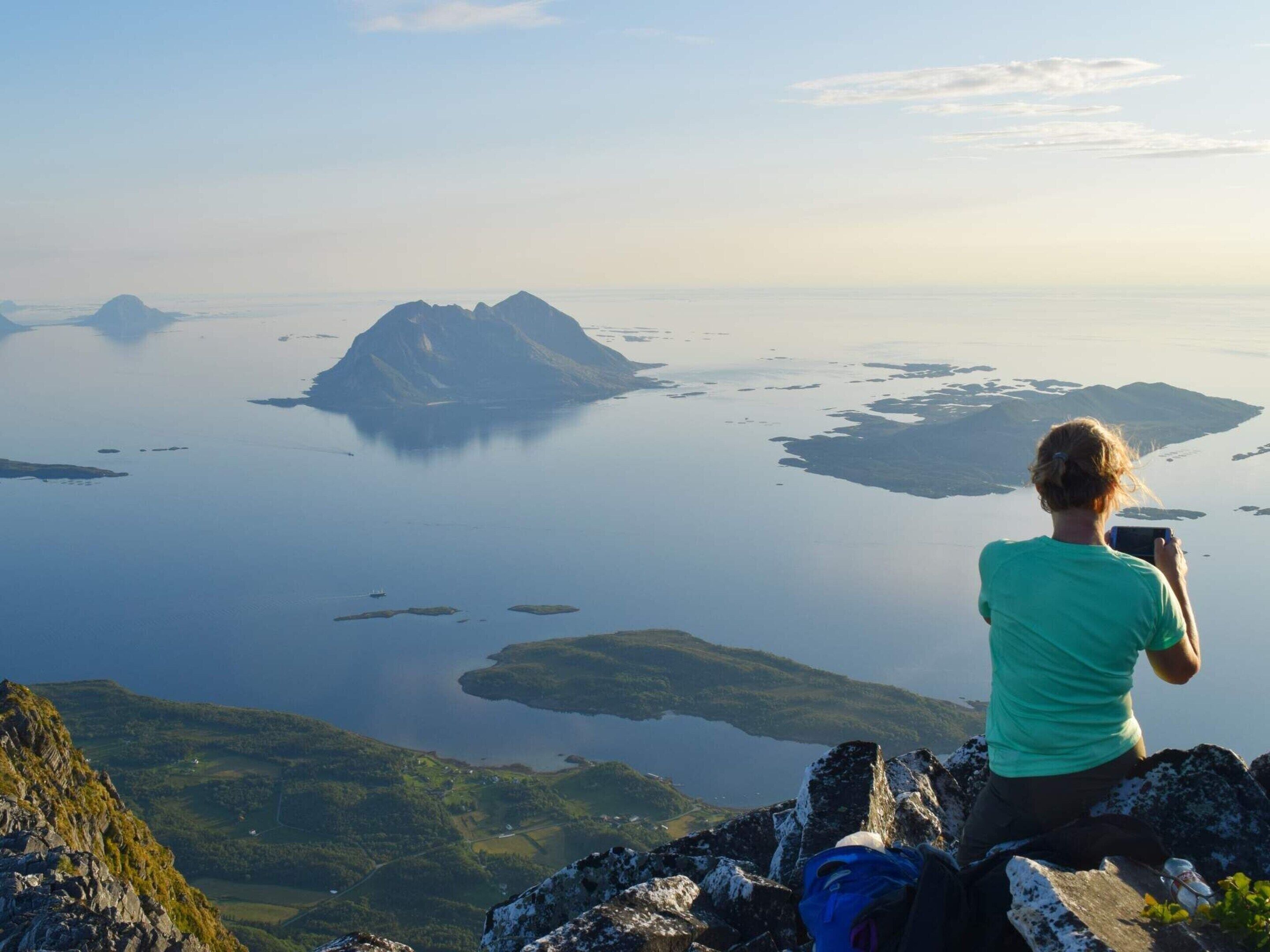 Eine Person sitzt auf einer Felsvorsprung und blickt auf einen ruhigen See mit mehreren Inseln, wobei sie die Aussicht mit einer Kamera festhält.