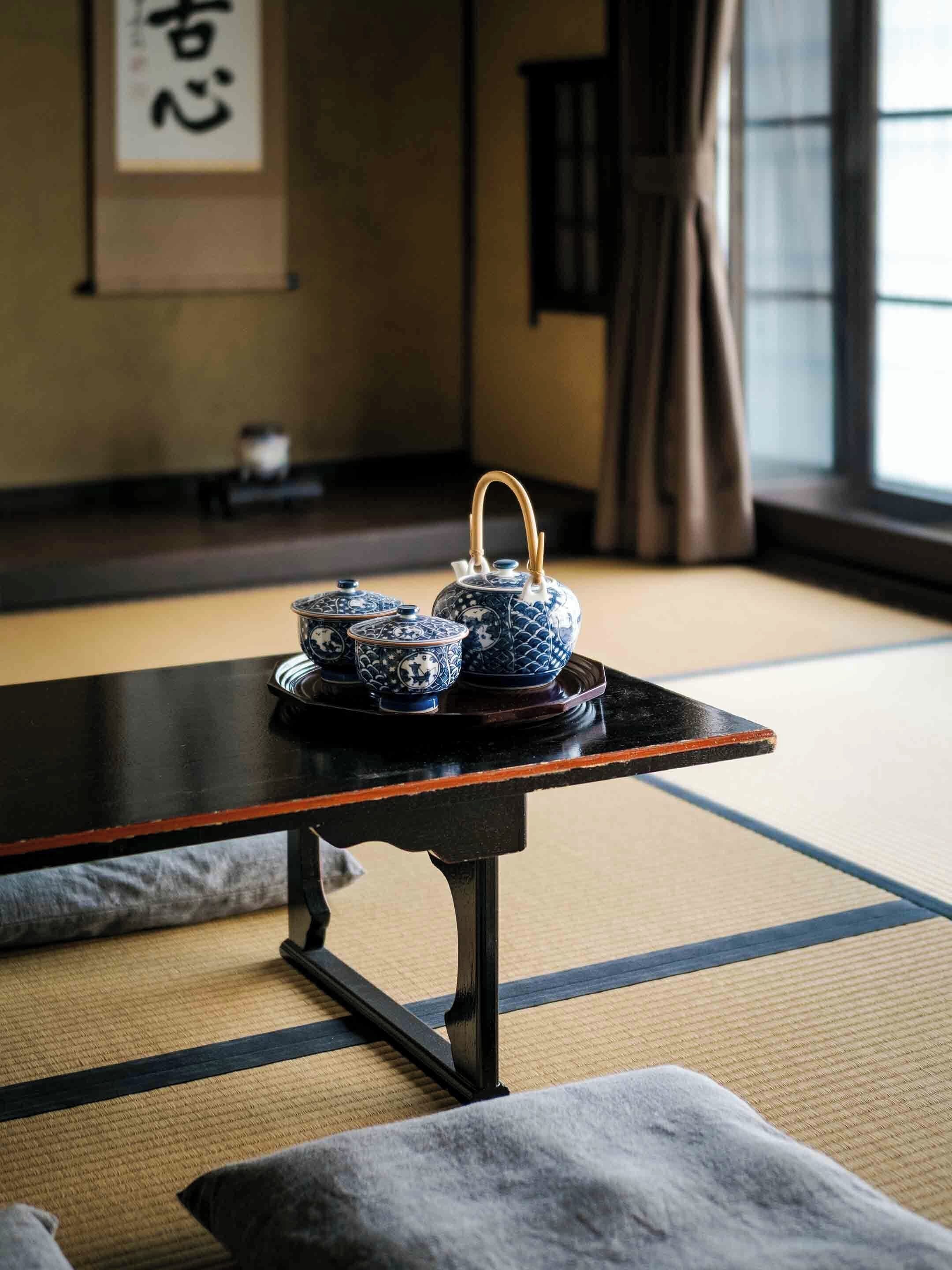 A tatami mat room with a black table holding three blue and white ceramic teapots, a framed calligraphy piece on the wall, and a window with curtains.