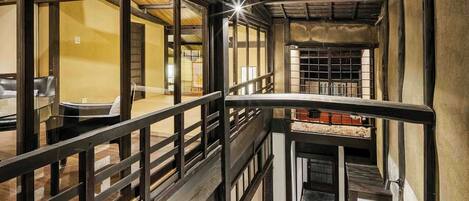 A narrow corridor with wooden railings and a stone-tiled floor, leading to a dining area with tables and chairs.