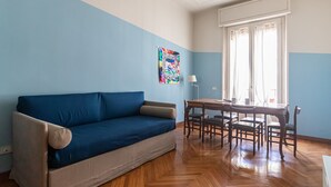 A room with a blue sofa, a wooden dining table with chairs, and a window with curtains. - Italianway Comfort - Gessi 49 (Milan)
