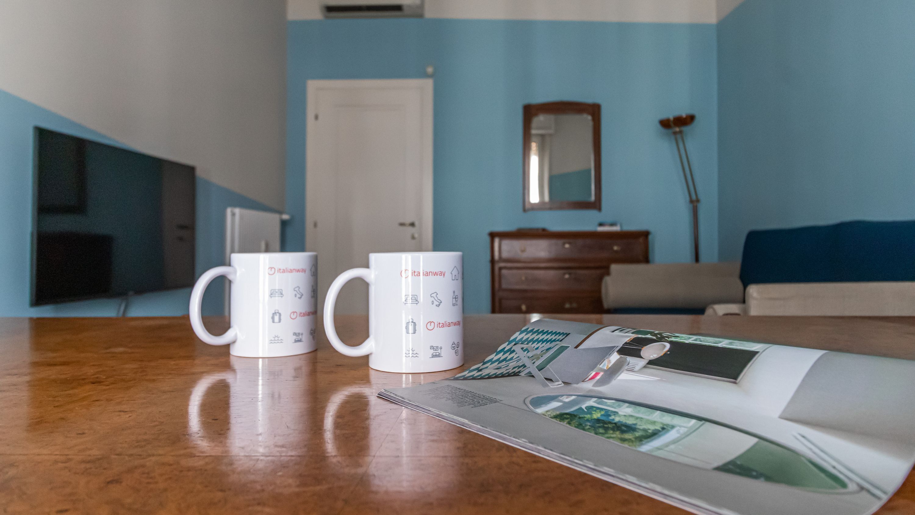 A room with a wooden table, two white mugs with Italianway branding, a magazine, a mirror, a dresser, and a blue wall.