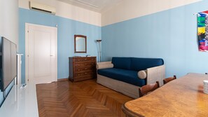 A room with a wooden herringbone floor, a blue sofa, a wooden dresser, a white door, a mirror, a lamp, and a dining table with chairs. - Italianway Comfort - Gessi 49 (Milan)