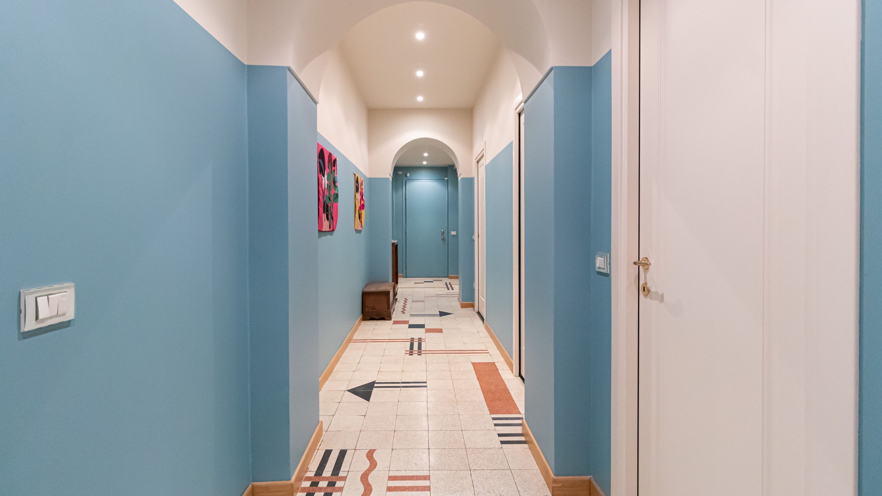 Um corredor com paredes azuis, chão de azulejos com setas indicativas e uma porta no final.