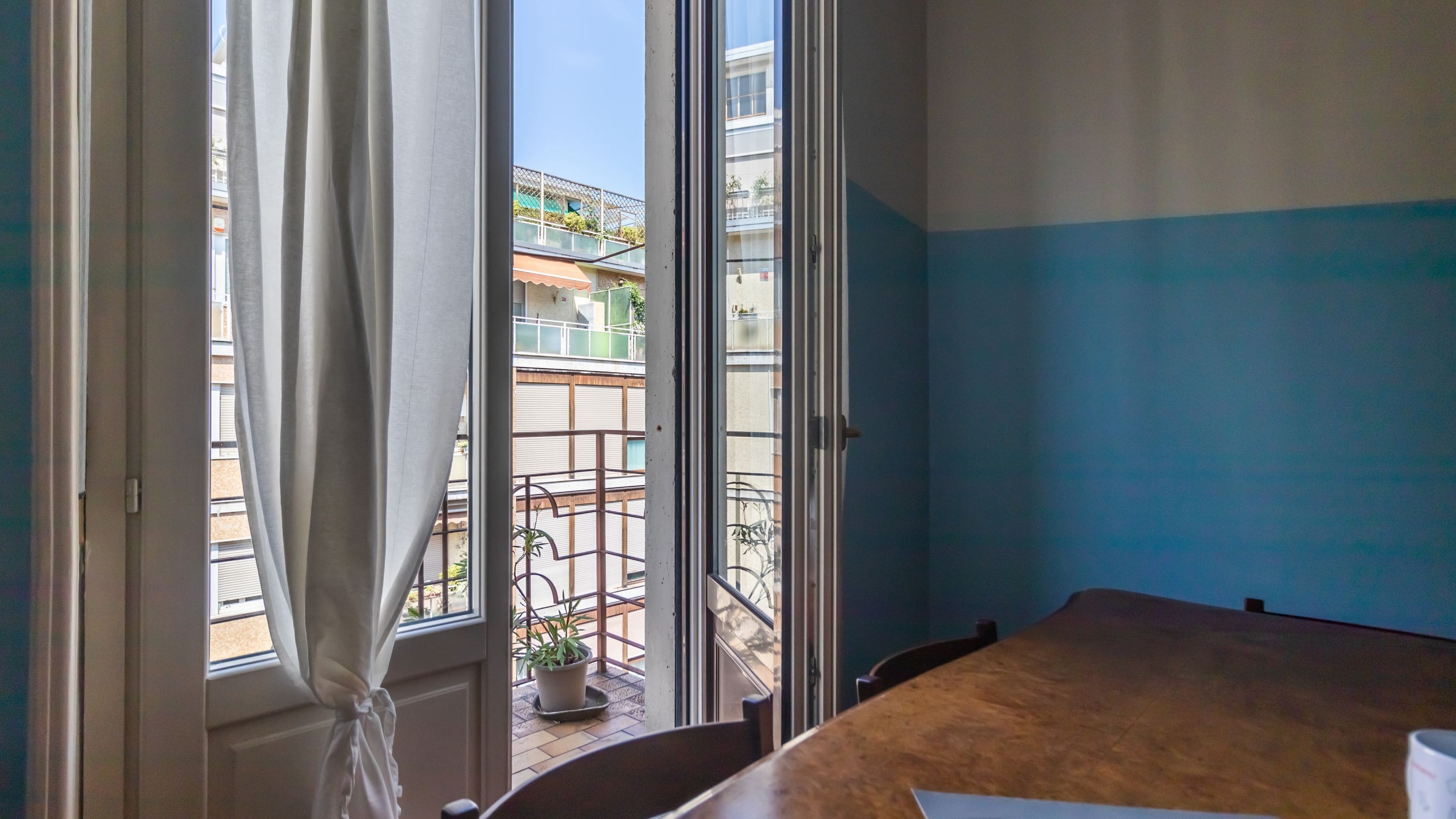 A room with a large window, a wooden table, and a view of a balcony with a railing and potted plants.