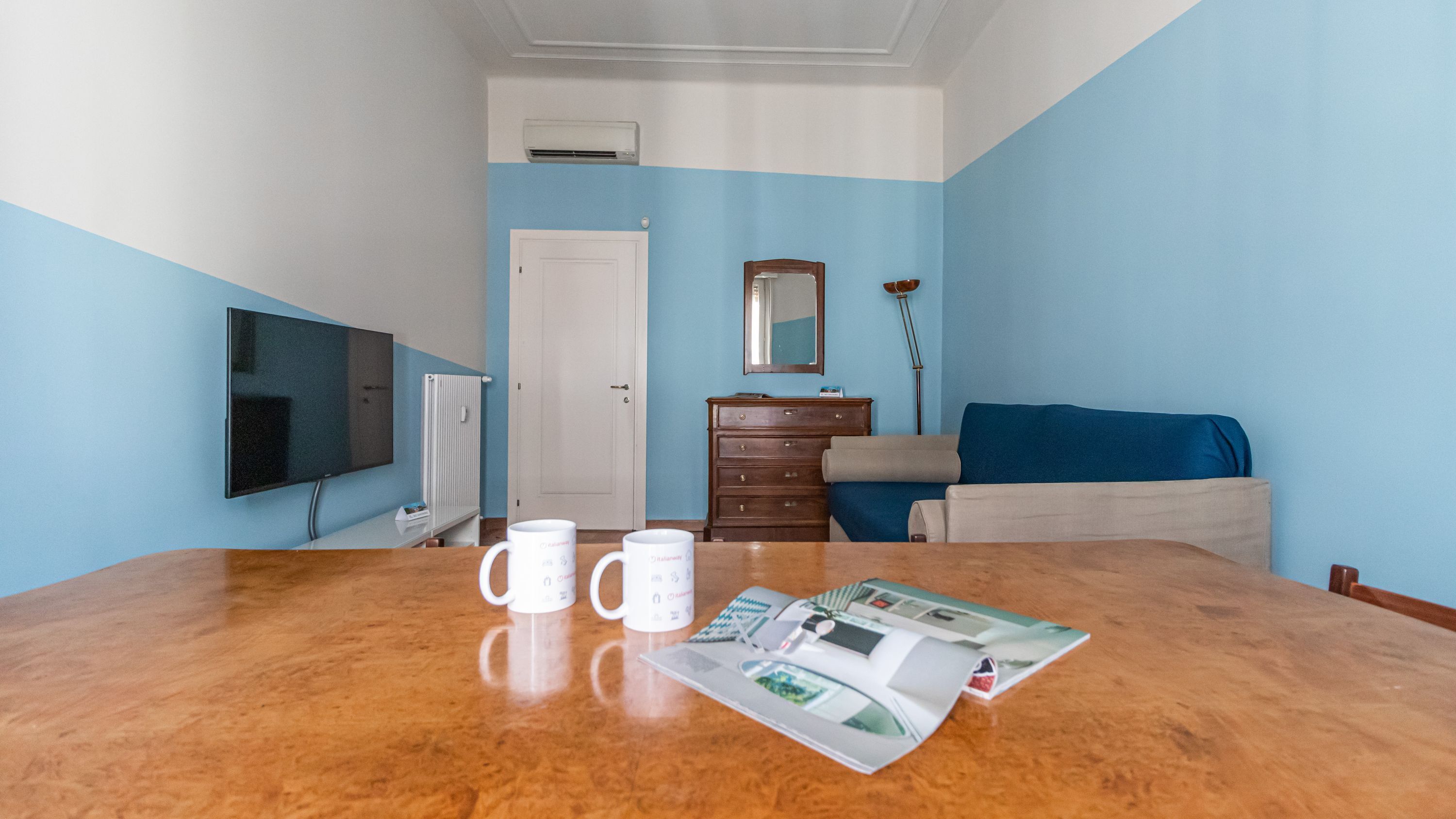 A room with a wooden table, two white mugs, a magazine, a dresser, a blue sofa, a TV, and a door.