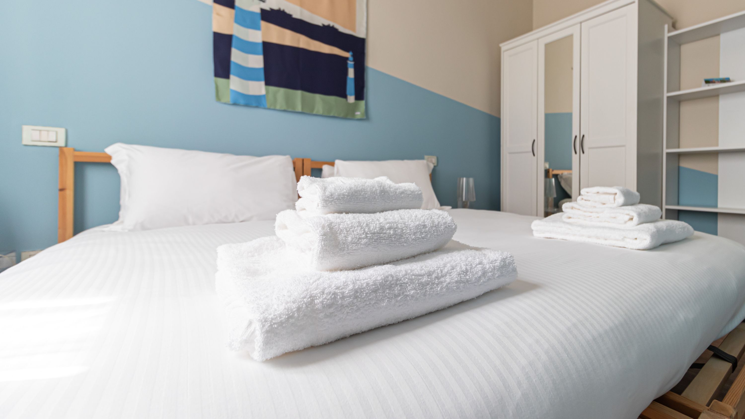 A neatly made bed with white linens and towels, a wooden headboard, a blue wall with a painting, and a white wardrobe with doors.