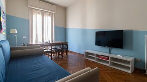 A living room with a blue sofa, a dining table with chairs, a flat-screen TV mounted on the wall, and a white entertainment unit with shelves. - Italianway Comfort - Gessi 49 (Milan)