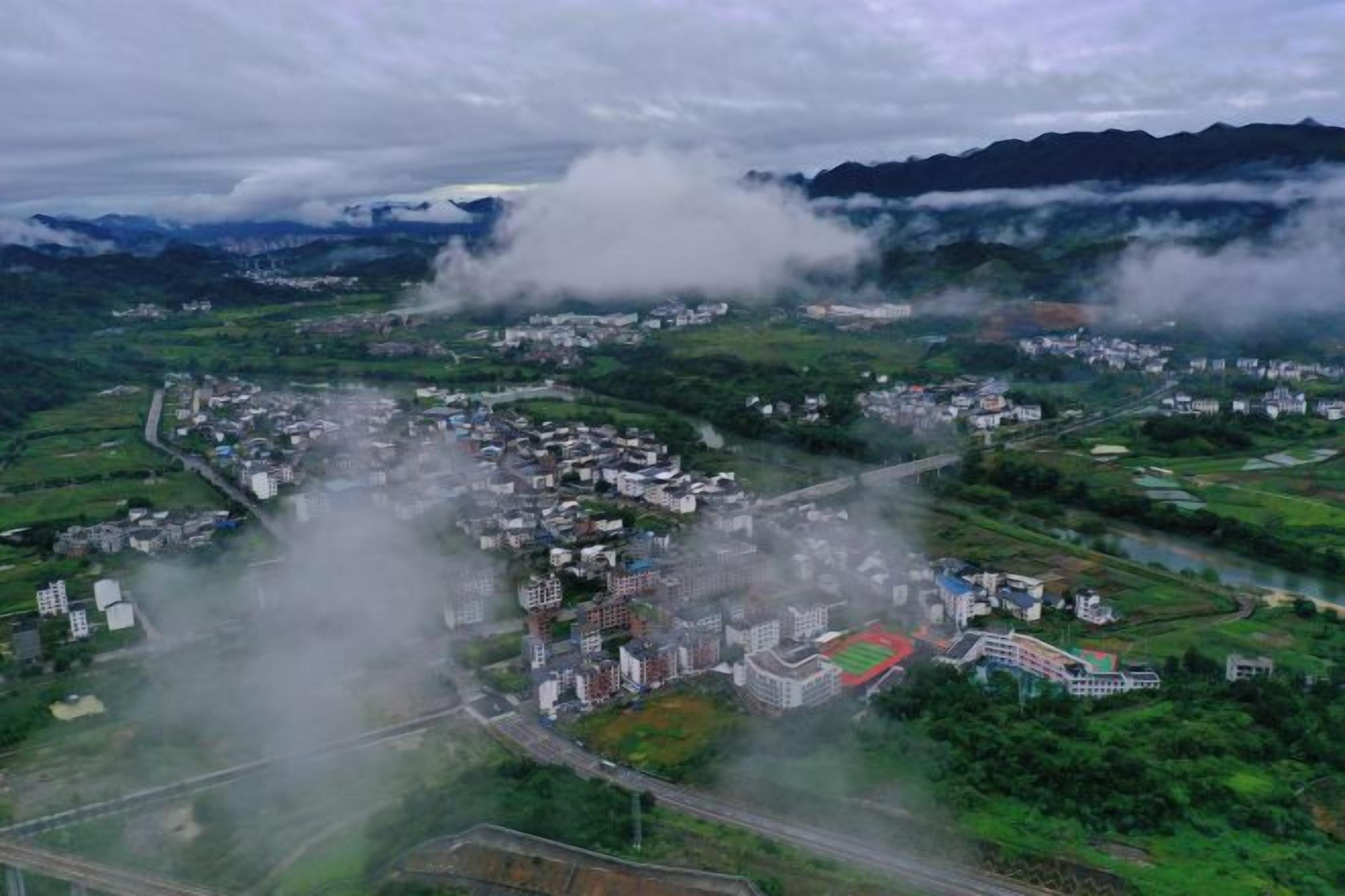 An aerial view of a town with a dense cluster of buildings, a river, and a mountainous backdrop.