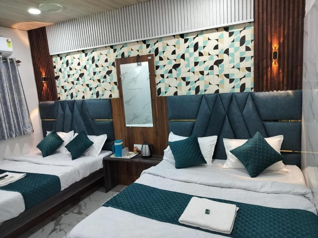 A hotel room with two beds, each with a dark headboard, white bedding, and teal throw pillows. A wooden nightstand with a coffee maker and a small table with a lamp is between the beds.
