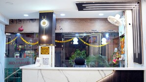 A hotel reception area with a marble counter, a reception desk, a glass door, and a wall with various decorations and a sign indicating check-in times. - Hotel Mahalok Palace (Ujjain)