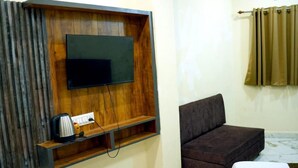 A room with a wooden TV stand, a flat-screen TV, a sofa, and a curtain. - Hotel Mahalok Palace (Ujjain)
