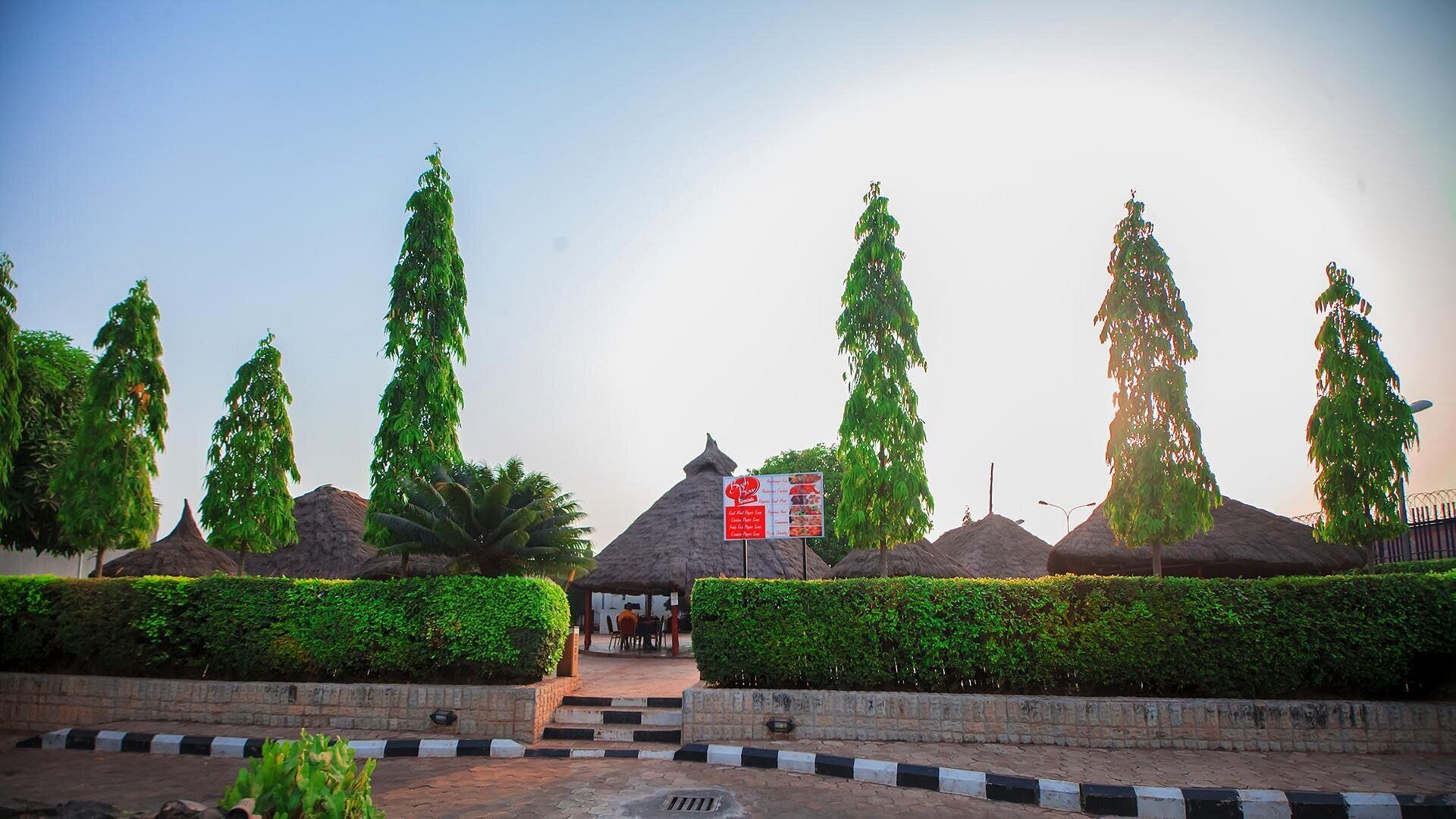 A well-maintained garden with neatly trimmed hedges and tall trees, a thatched-roof structure, and a signboard likely indicating a restaurant or cafe.