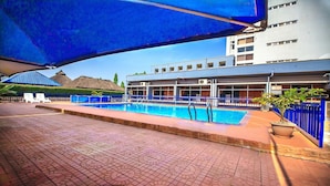 A swimming pool with a tiled deck, a covered area with blue canopies, and a building with multiple windows and balconies in the background. - Universal Hotel Enugu (Enugu)