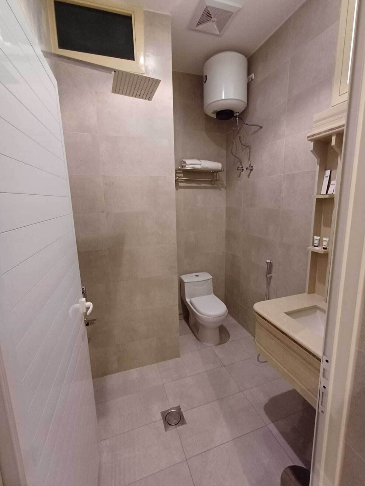 Een badkamer met toilet, wastafel en douche. Boven het toilet hangt een handdoekhouder en er is een plank met toiletartikelen.