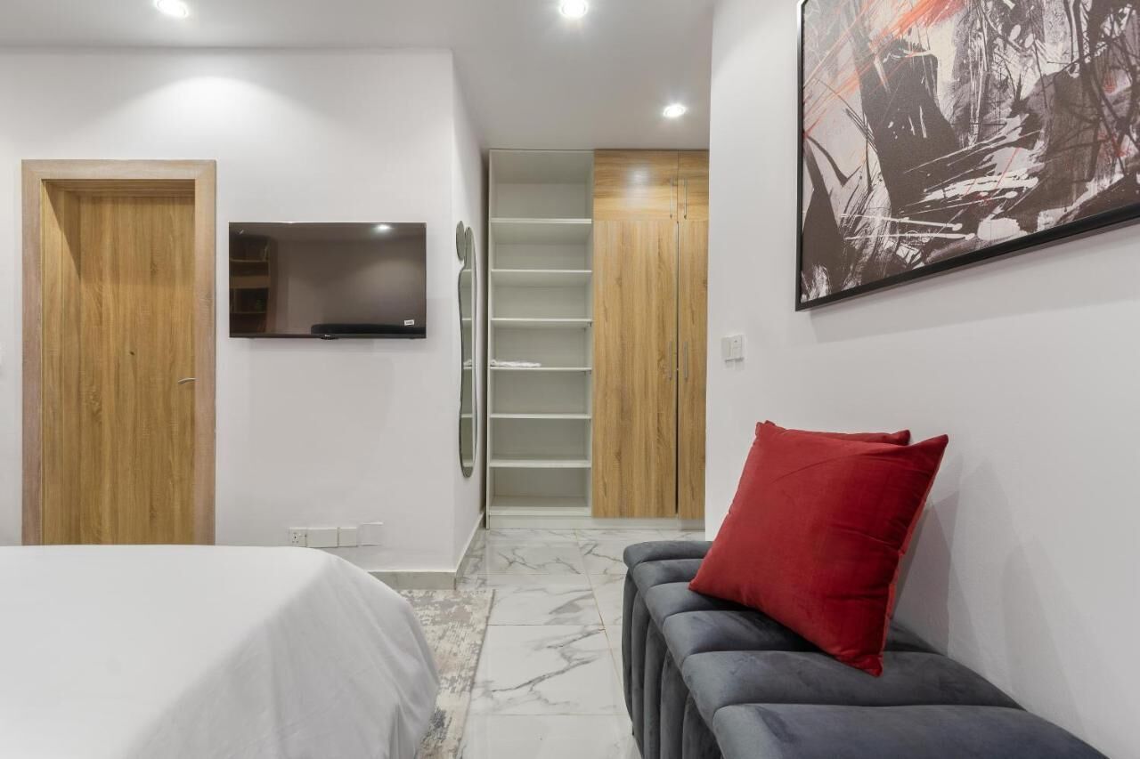 Ein modernes Schlafzimmer mit Bett, grauem Sofa mit rotem Kissen, Wandfernseher und Holzschrank.