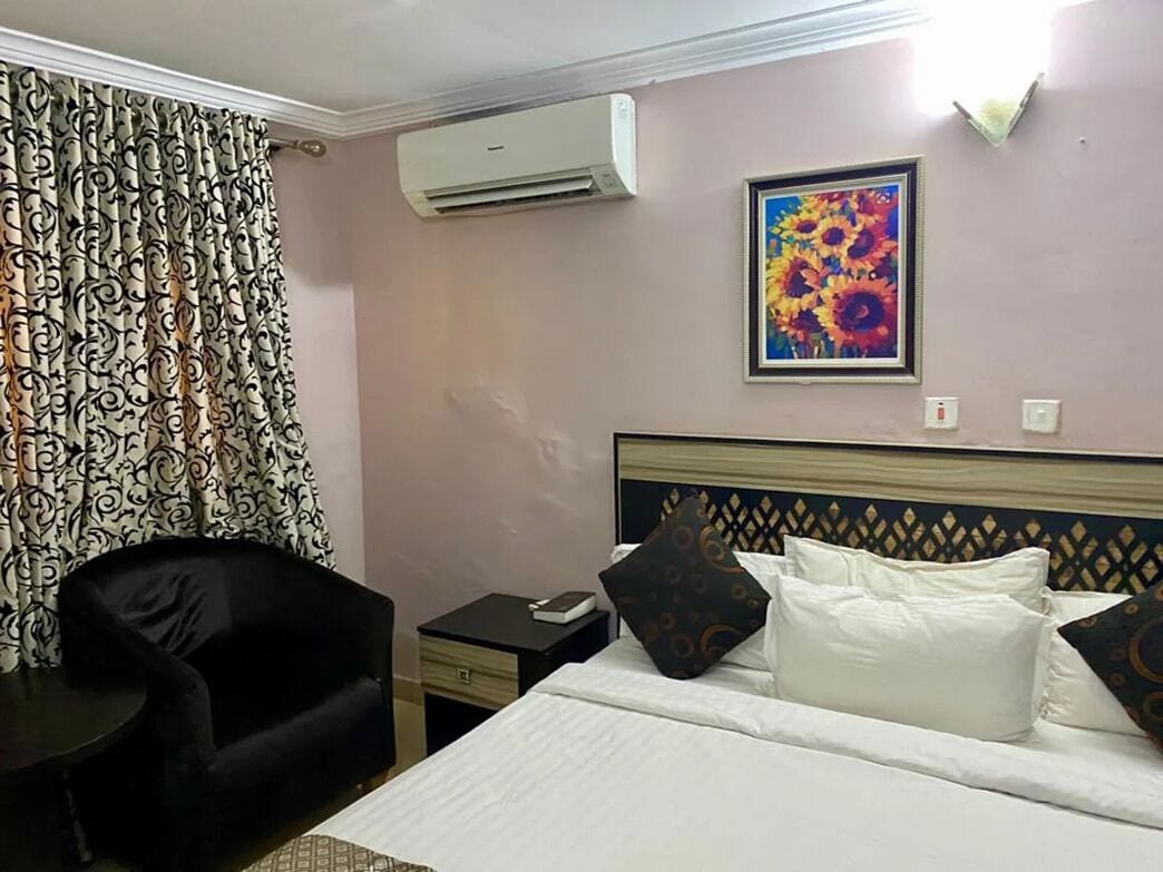 A hotel room with a bed, a chair, a nightstand, an air conditioner, and a painting on the wall.