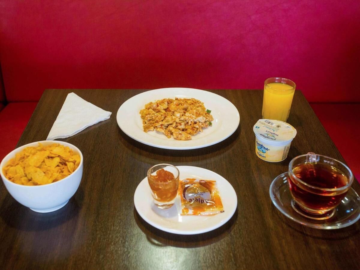 Une table avec une assiette de nourriture, un bol de céréales, un verre de jus d’orange, une tasse de thé et un petit pot de yaourt.