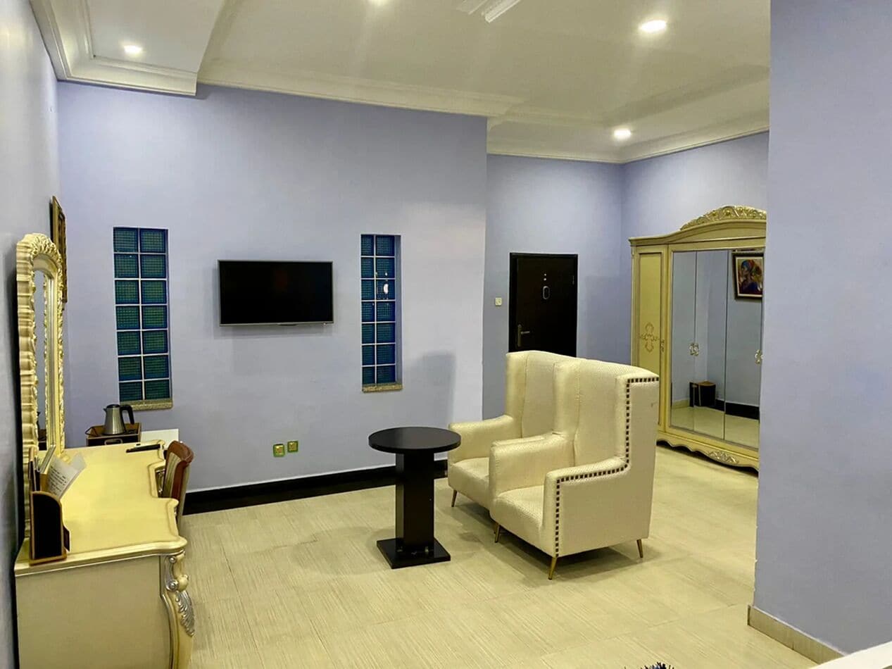 A room with a flat-screen TV, a cream-colored armchair, a small round table, and a mirror with a decorative frame.