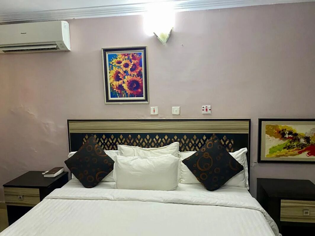 A hotel room with a bed, two bedside tables, an air conditioning unit, and two wall-mounted paintings.