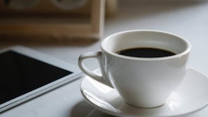 A white cup of coffee on a saucer next to a tablet on a table. - Garden Atlantis Hotel (Enugu)