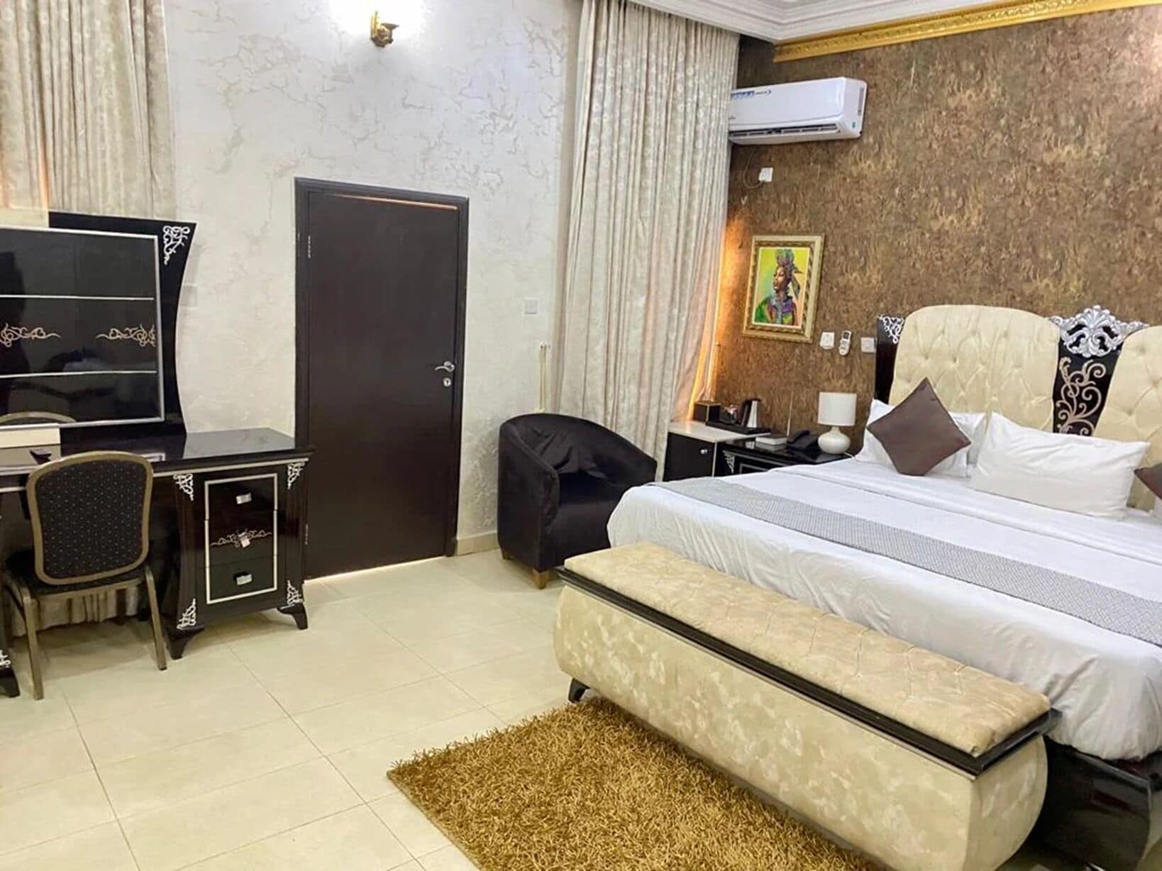 A hotel room with a bed, a desk with a chair, a television, a wardrobe, and an air conditioning unit.