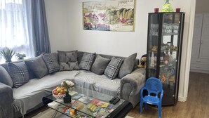 A modern living room with a grey sectional sofa, a glass coffee table with magazines and snacks, a blue chair, a glass cabinet with decorative items, and a traditional painting on the wall. - Cosy London Apartment Near Borough Market (London)