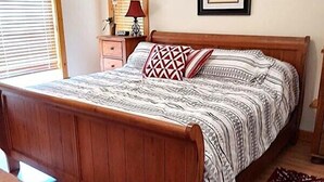 A bedroom with a wooden bed, a nightstand, a lamp, a framed picture on the wall, and a window with blinds.