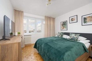 Une chambre à coucher avec un lit, un téléviseur posé sur un meuble en bois, une fenêtre avec des rideaux et des tableaux encadrés accrochés au mur.