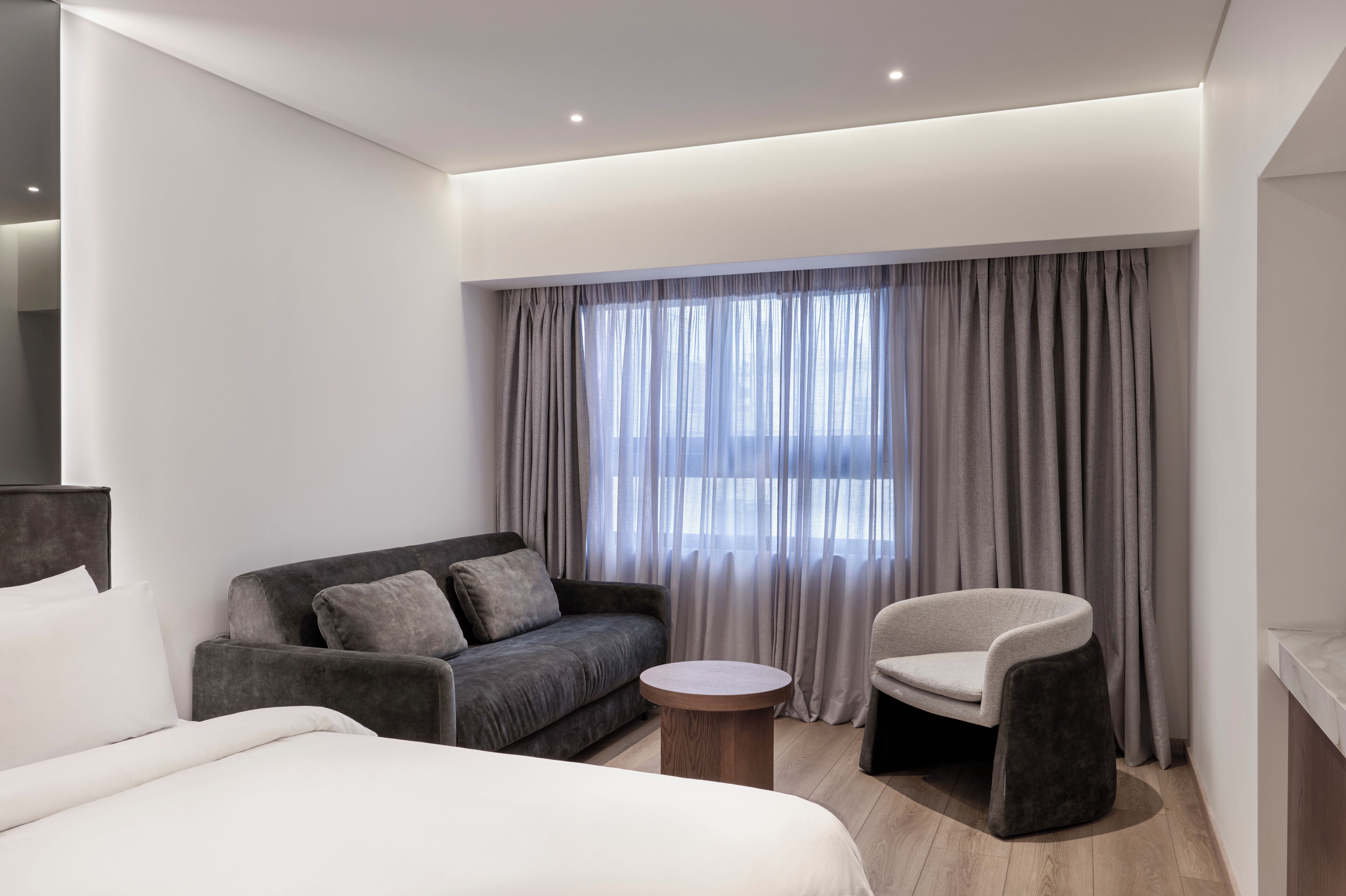 A modern hotel room with a bed, a grey sofa, a round wooden side table, and a chair.
