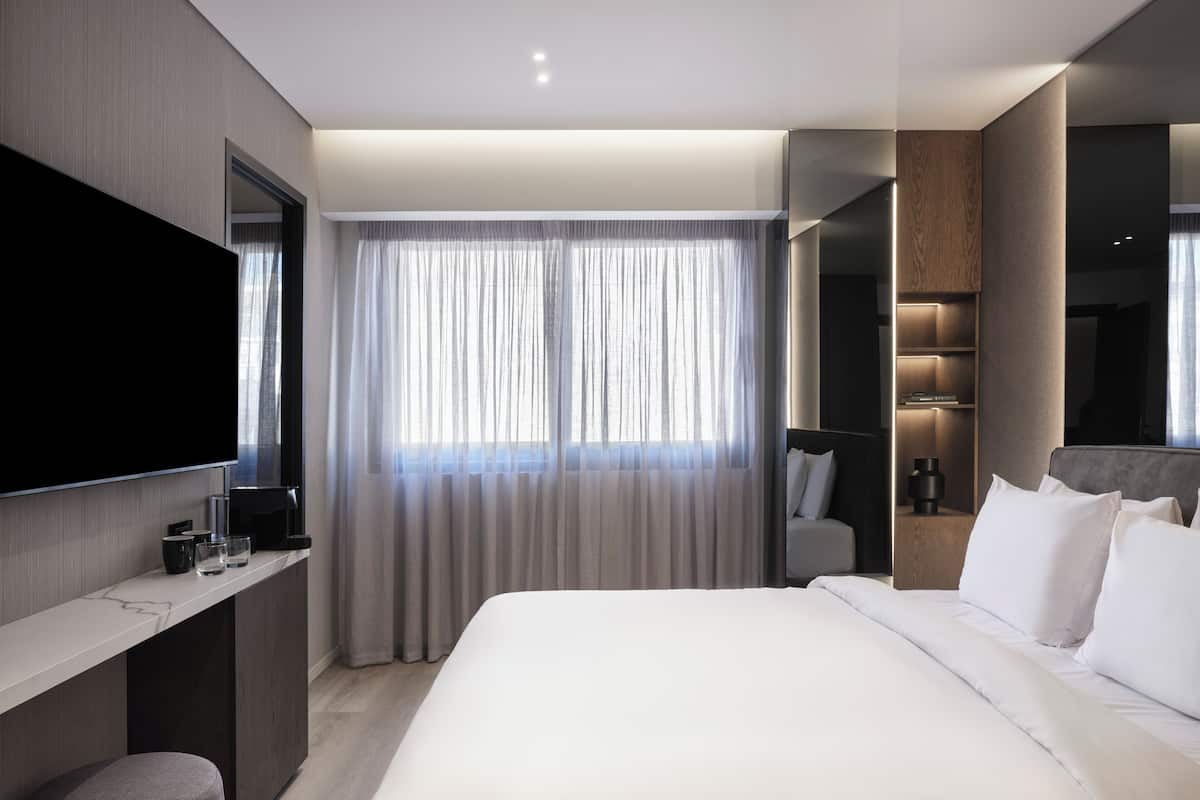 A modern hotel room with a large bed, a flat-screen TV mounted on the wall, a bedside table with a lamp, and a window with sheer curtains.