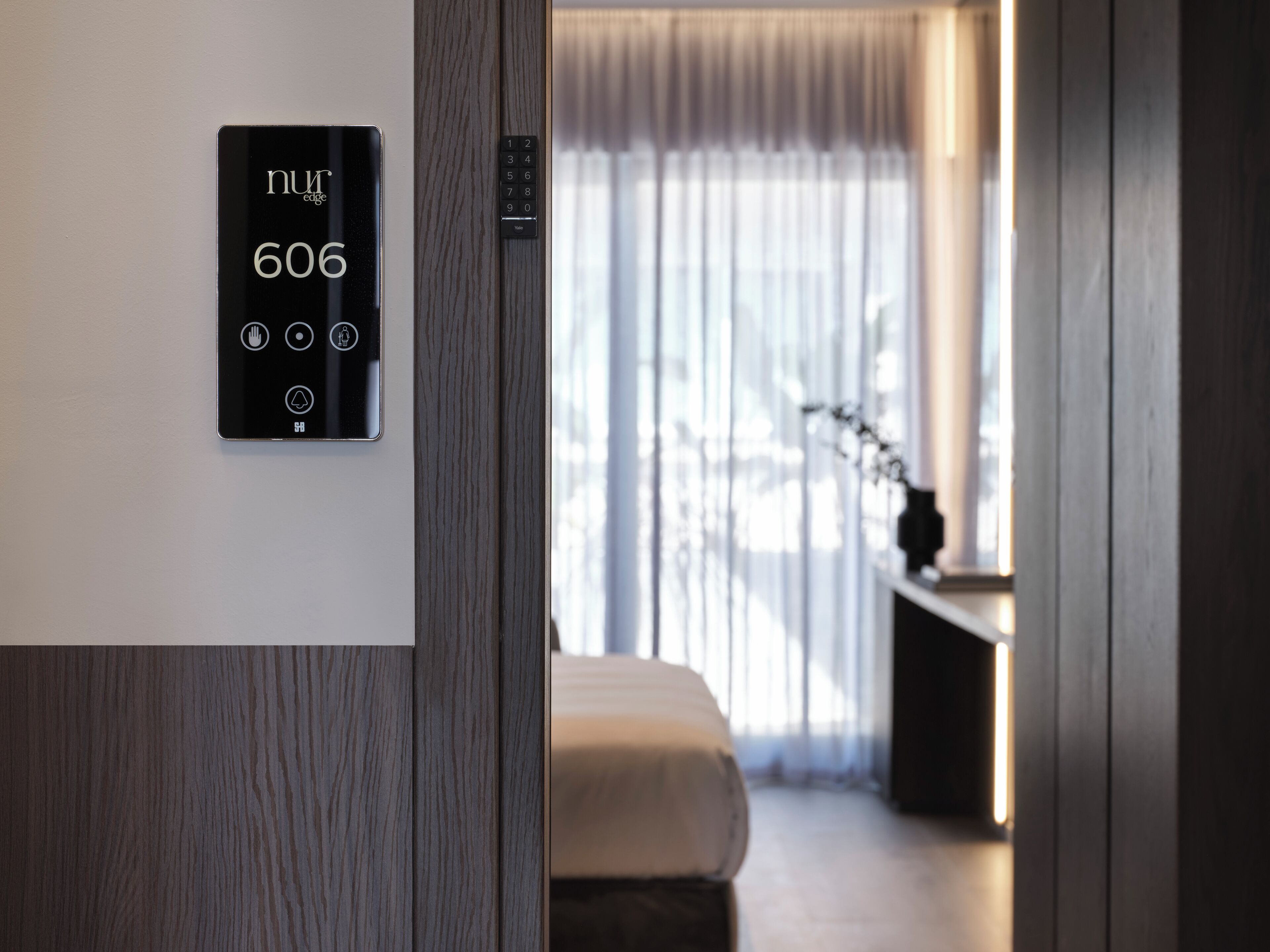 A modern hotel room with a digital keypad displaying room number 606, a wooden panel wall, a bed, and a large window with curtains.