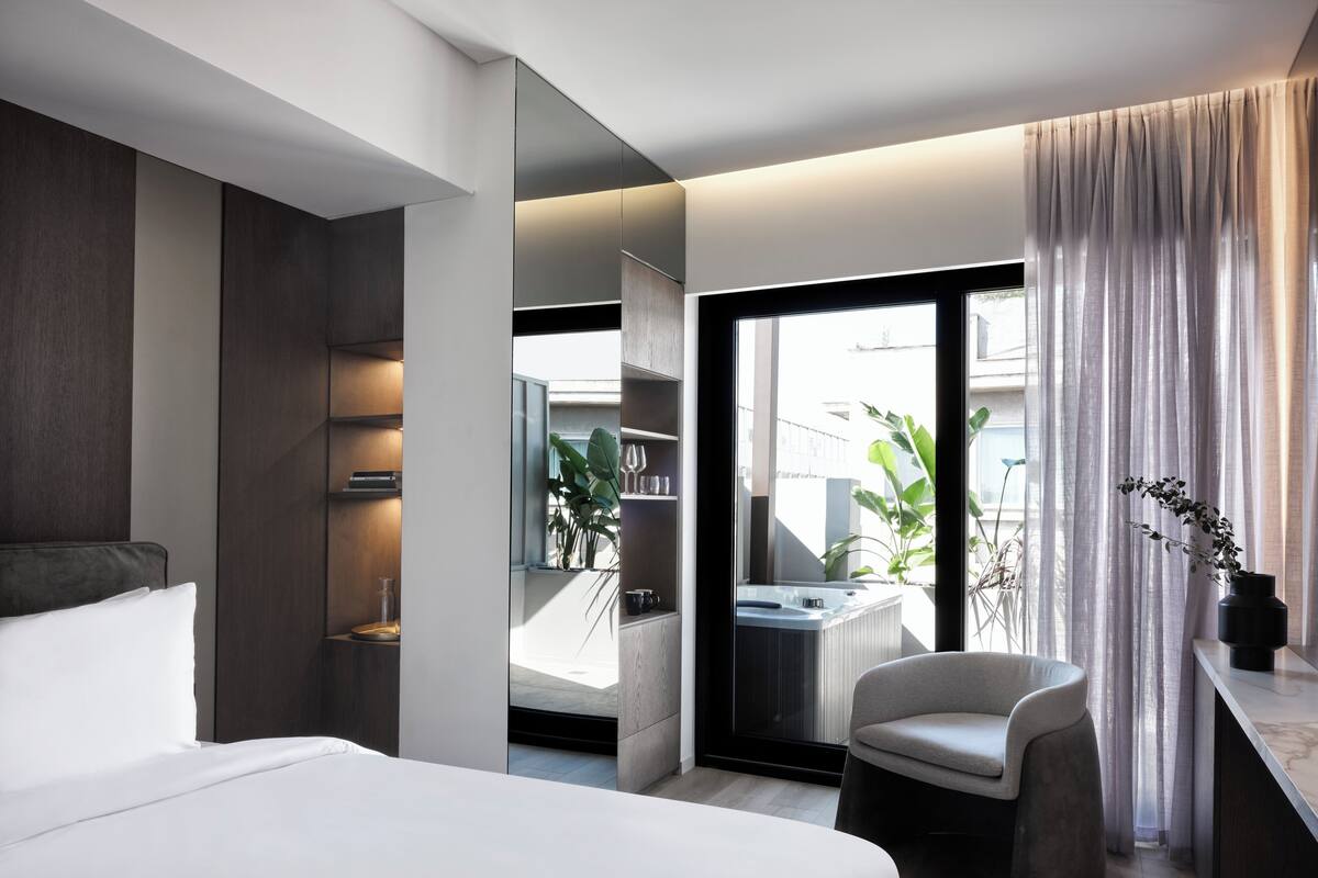 A modern hotel room with a large bed, a seating area, a view of the outdoors, and a bathroom with a bathtub.