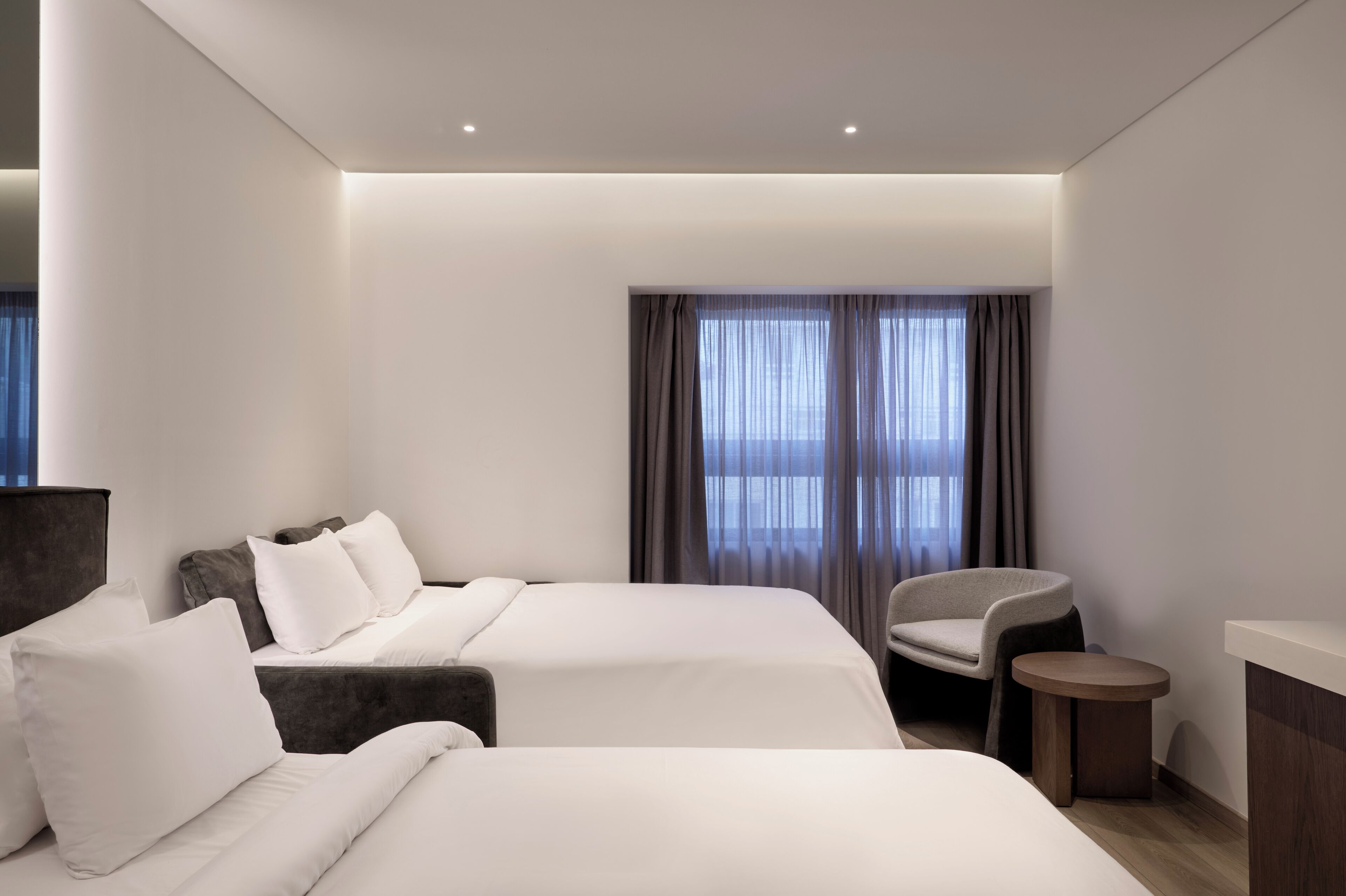 A modern hotel room with two beds, a chair, a small table, and a large window with curtains.