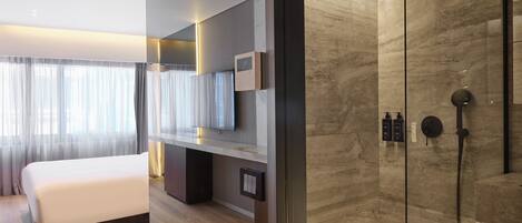 A modern hotel room with a large bed, a TV mounted on the wall, a digital clock, and a shower area with a glass enclosure.