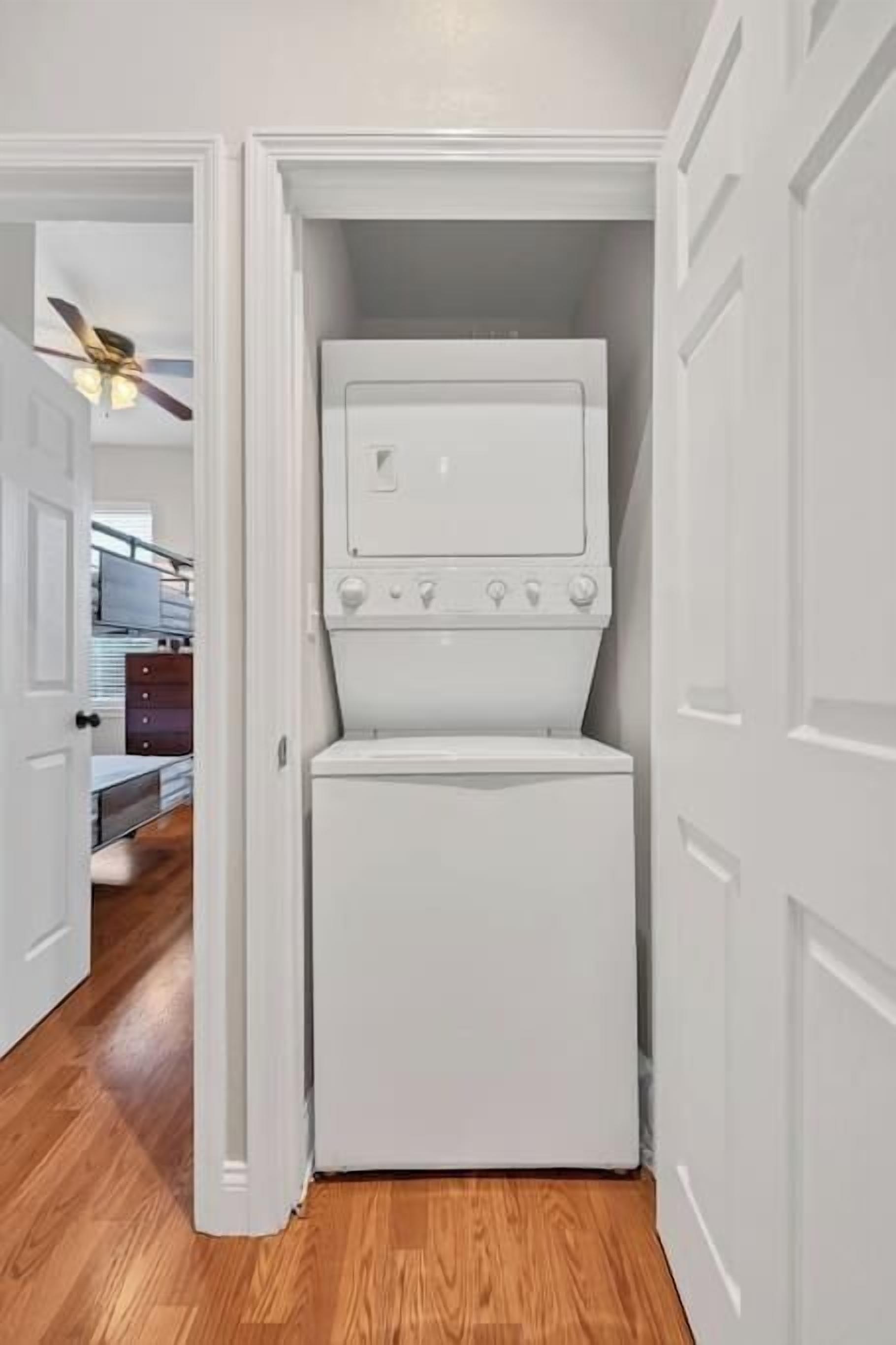 A compact laundry room with a front-loading washer and dryer, wooden flooring, and a door leading to a hallway with a ceiling fan.