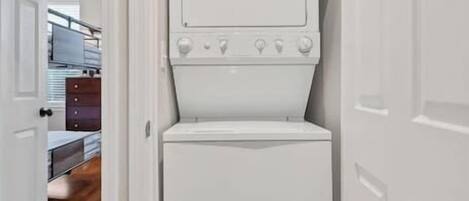 A compact laundry room with a front-loading washer and dryer, wooden flooring, and a door leading to a hallway with a ceiling fan.