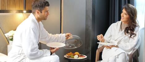 A couple in a hotel room, seated on a bed with a breakfast tray between them.