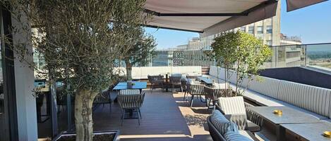 An outdoor patio area with seating and tables, surrounded by olive trees and a view of the cityscape.
