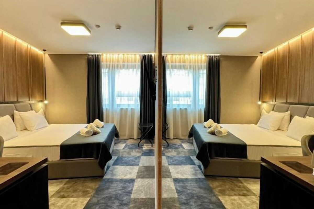 Habitación de hotel con espejo, dos camas, suelo de cuadros y vistas de la ciudad a través de amplios ventanales.
