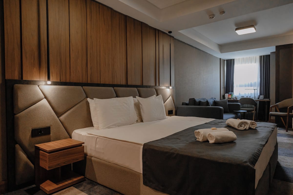 A modern hotel room with a large bed, a wooden headboard, a small bedside table, and a view of the city through the window.