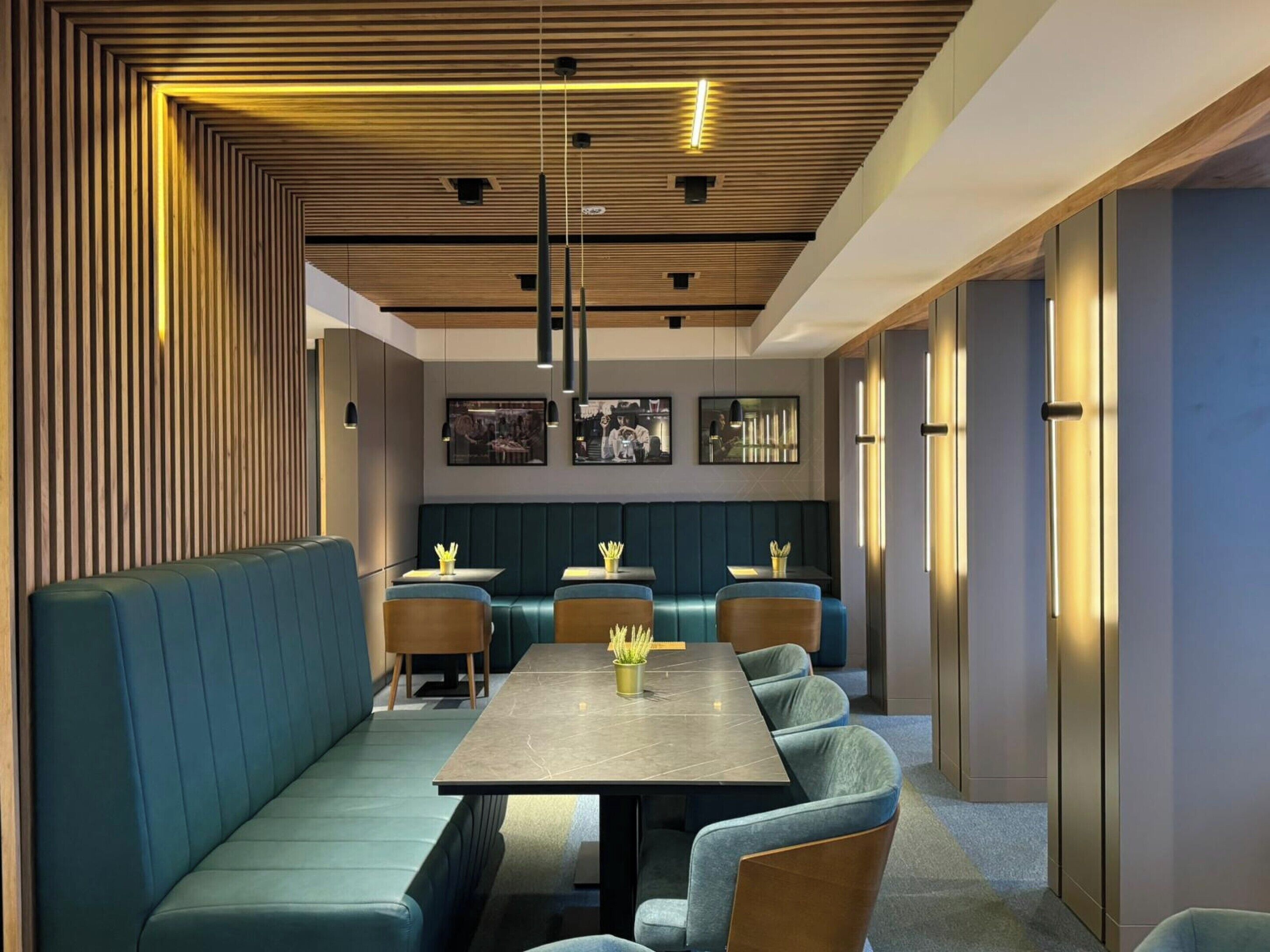 A modern restaurant with a long communal table, cushioned seating, and a wooden slatted ceiling.