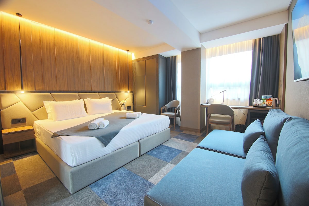 A modern hotel room with a large bed, a sofa, a desk, and a view of the sea.