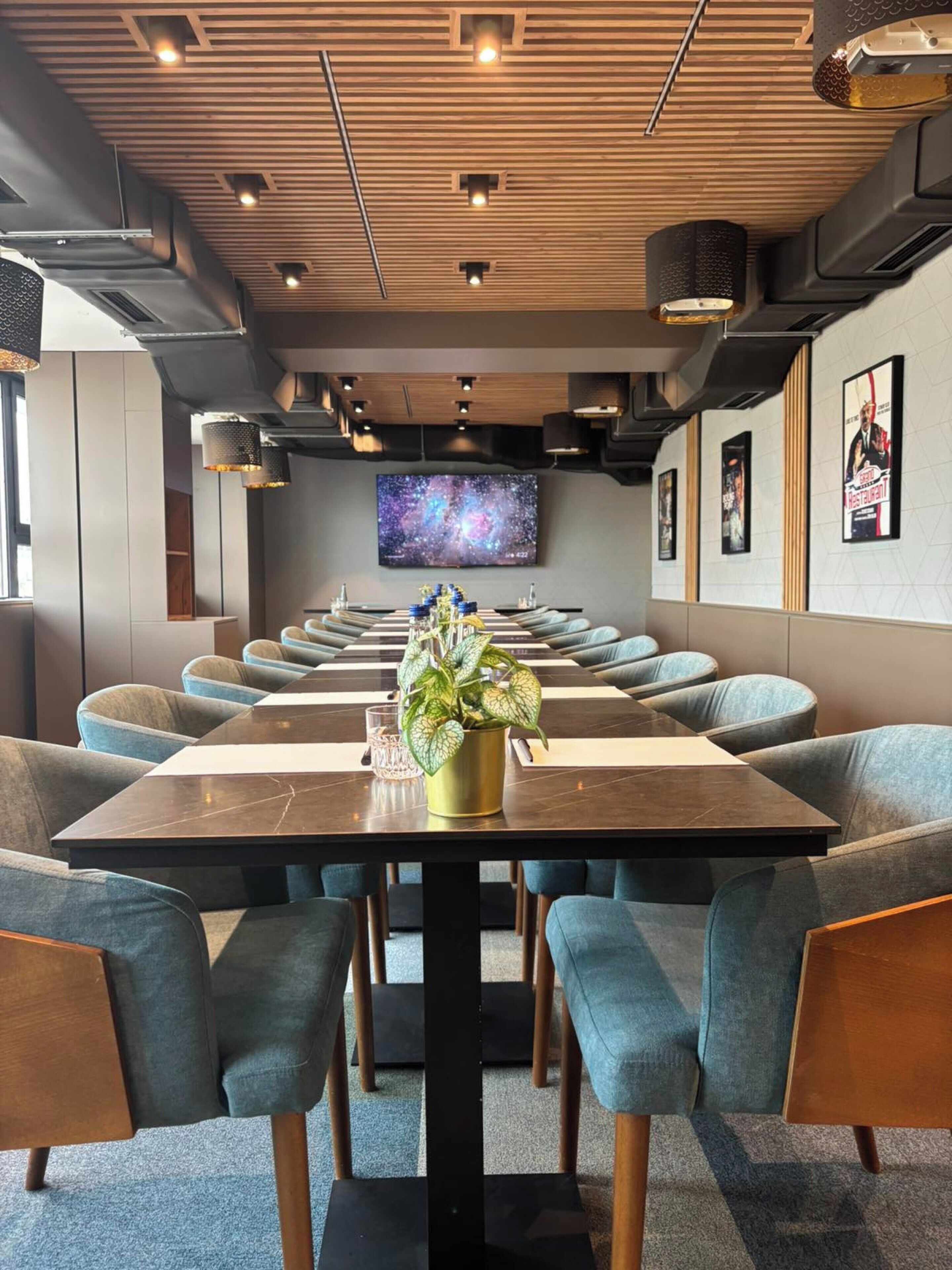 A modern conference room with a large table, blue upholstered chairs, and a television screen on the wall.