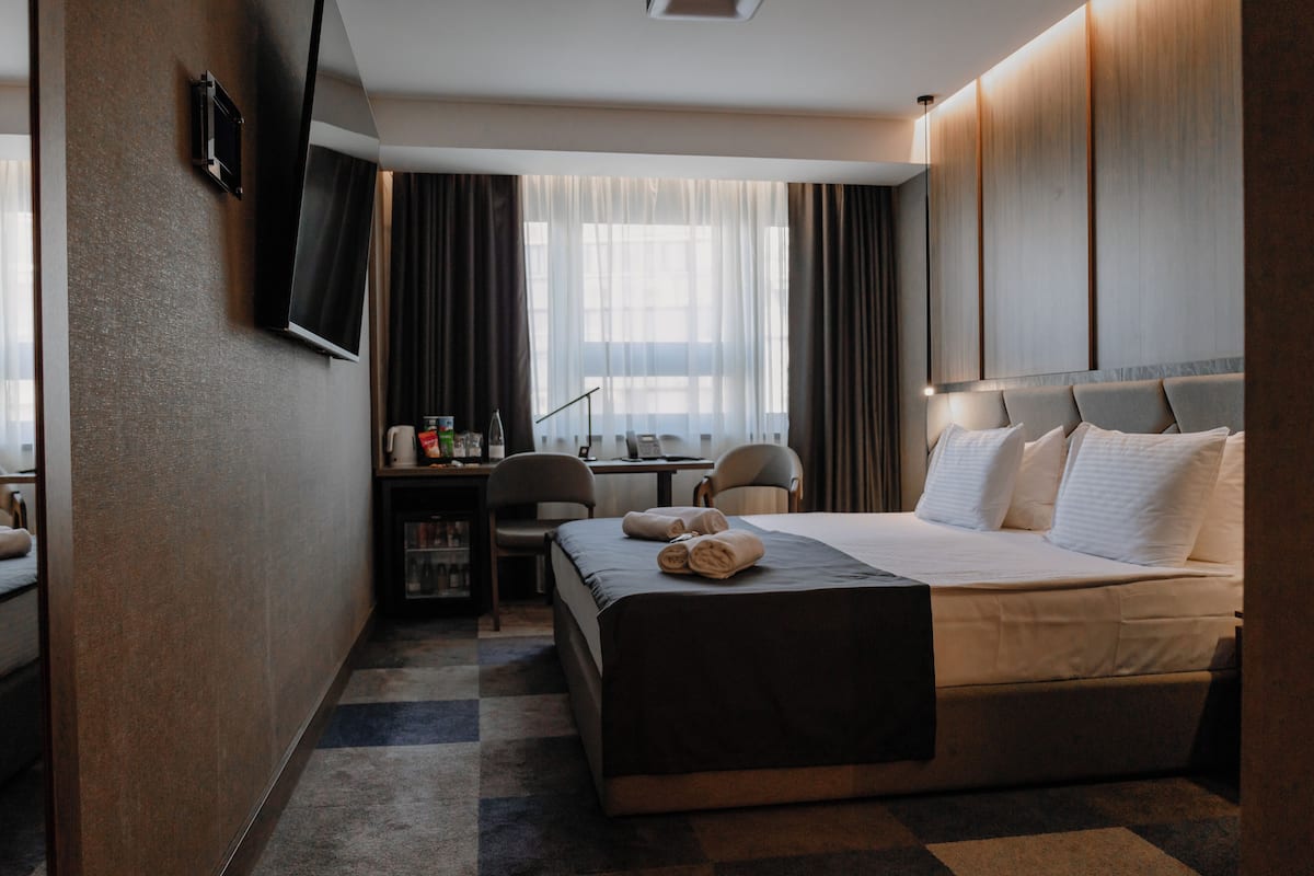 A hotel room with a large bed, a desk with a chair, a TV mounted on the wall, and a window with curtains.