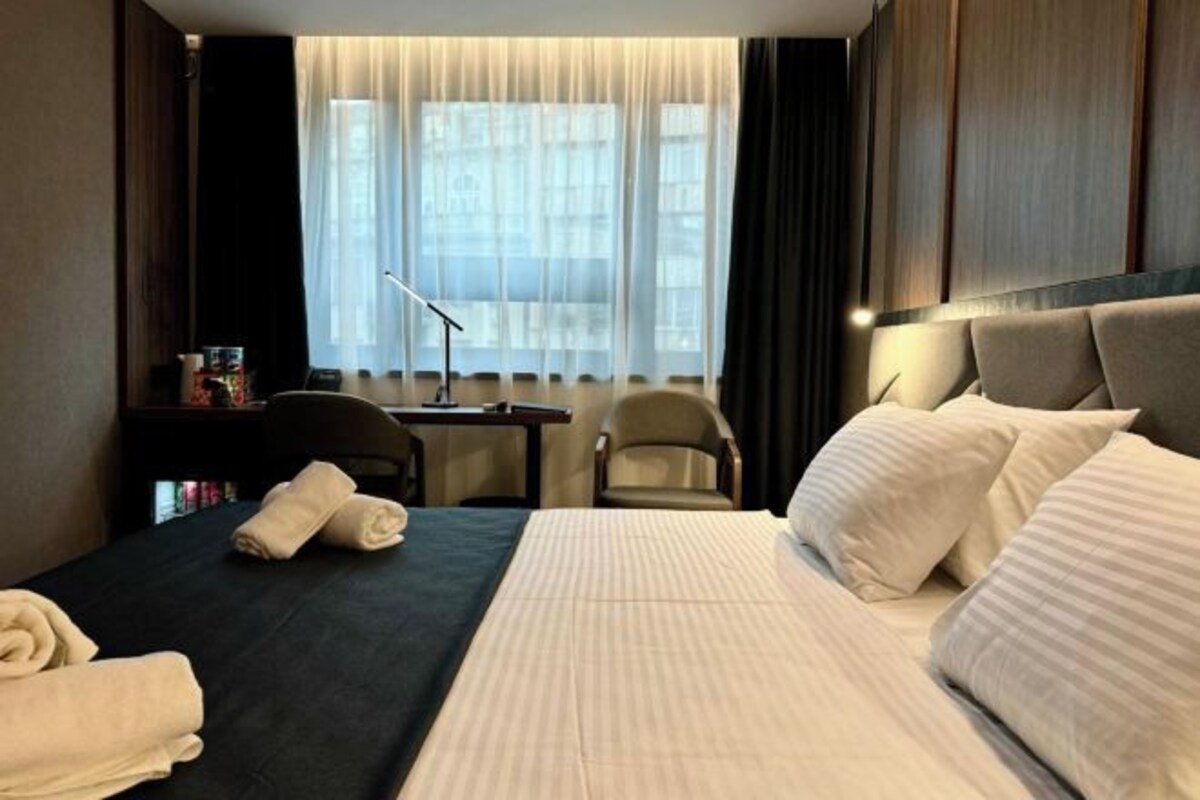 A hotel room with a large bed, a desk, a chair, and a window with sheer curtains.
