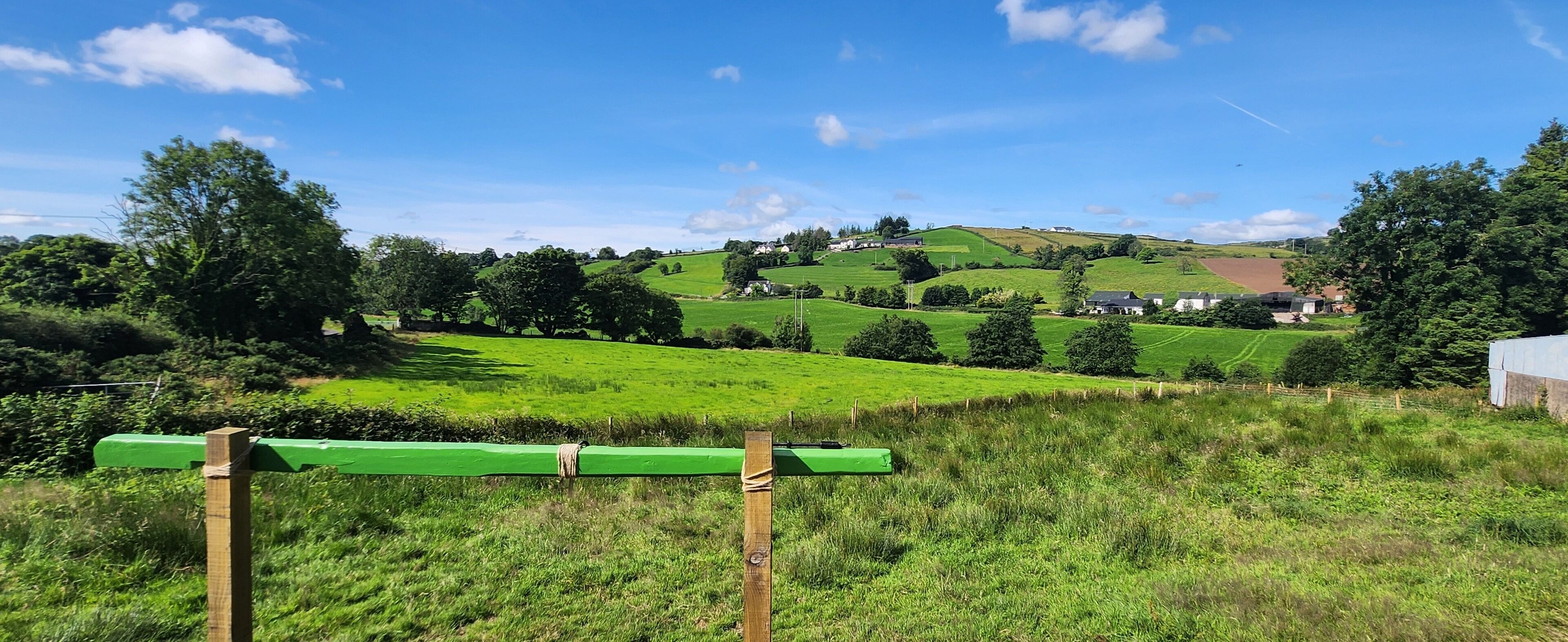 A green bench in a grassy field with a view of rolling hills and houses in the distance.
