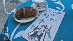 A table with a croissant and a cup of coffee, a menu, and a view of a street with buildings and a statue. - Heraklean Suites Tropea (Tropea)