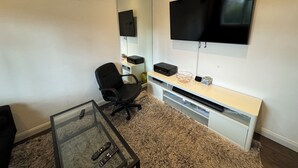 A modern office space with a large flat-screen TV mounted on the wall, a glass-top desk, a black office chair, and a carpeted floor.
