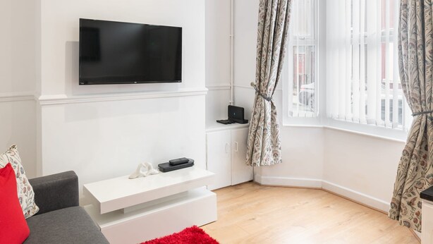 A modern living room with a flat-screen TV mounted on the wall, a white console table, a grey sofa with a red pillow, and a red rug. - Host Stay Bardsay Road (Liverpool)